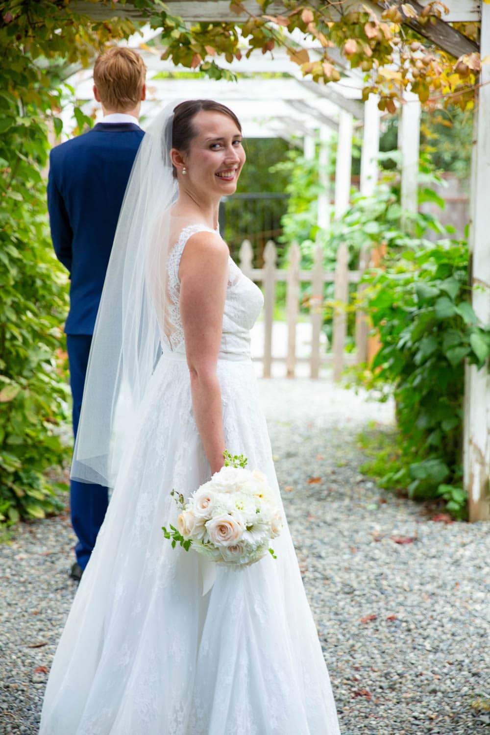 A bride turns and smiles at the camera. Her groom stands in front of her with his back to us because they are about to see each other for the first time on their wedding day.