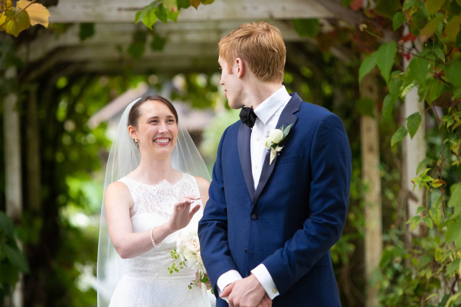 A groom in a navy suit turns to see his bride for the first time on their wedding day in this traditional "first look" photo. His bride stands behind him with her arm raised, smiling broadly at him.