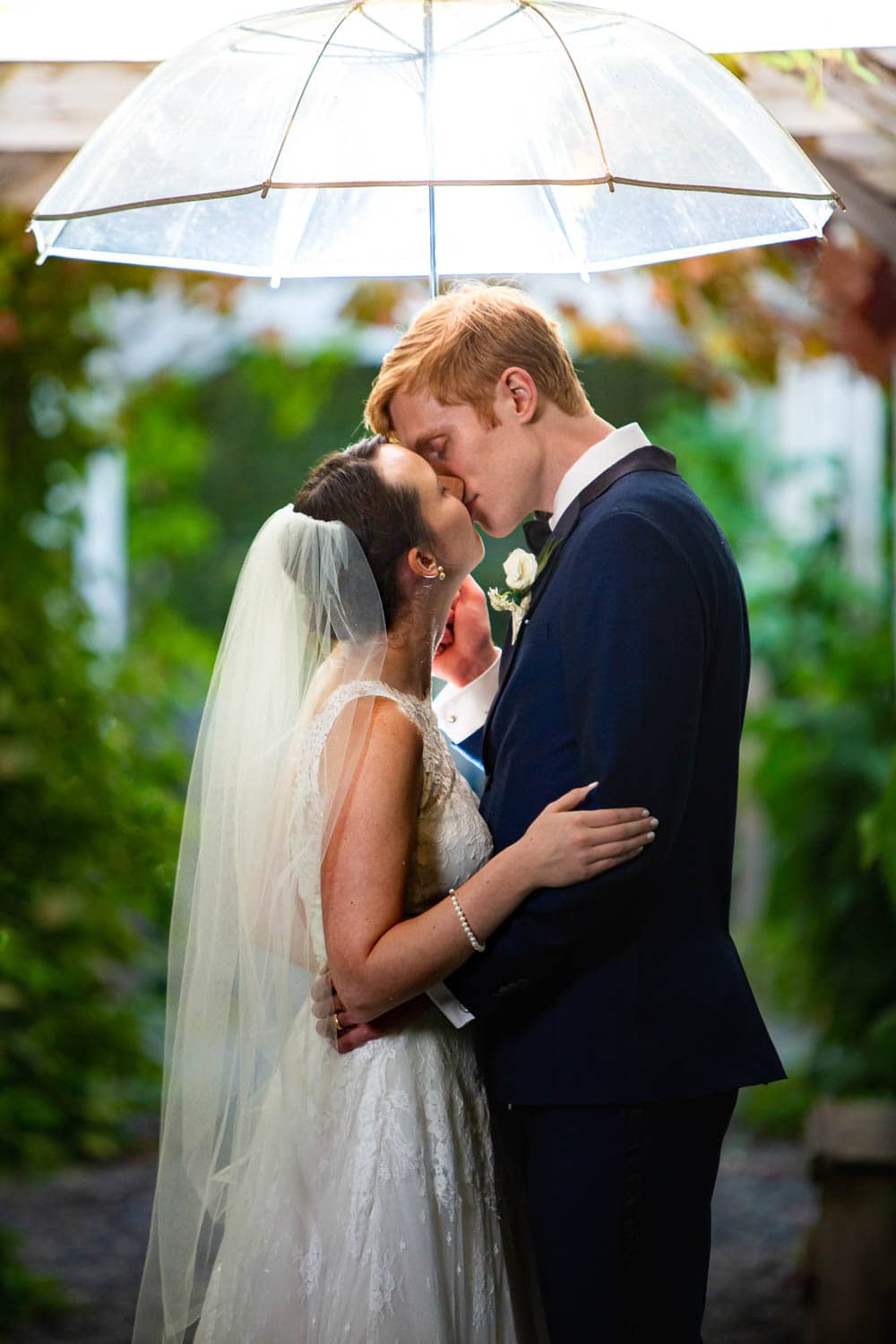 A bride and groom kiss under a clear umbrella in a lush area of Black Diamond Gardens.
