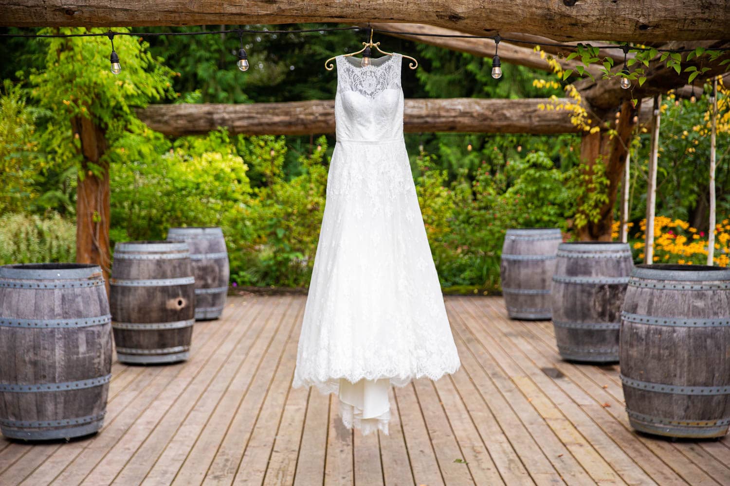 The bride's wedding dress hangs on a unique gold hangar with curled ends surrounded by a rustic outdoor arbor with wine barrel decor.