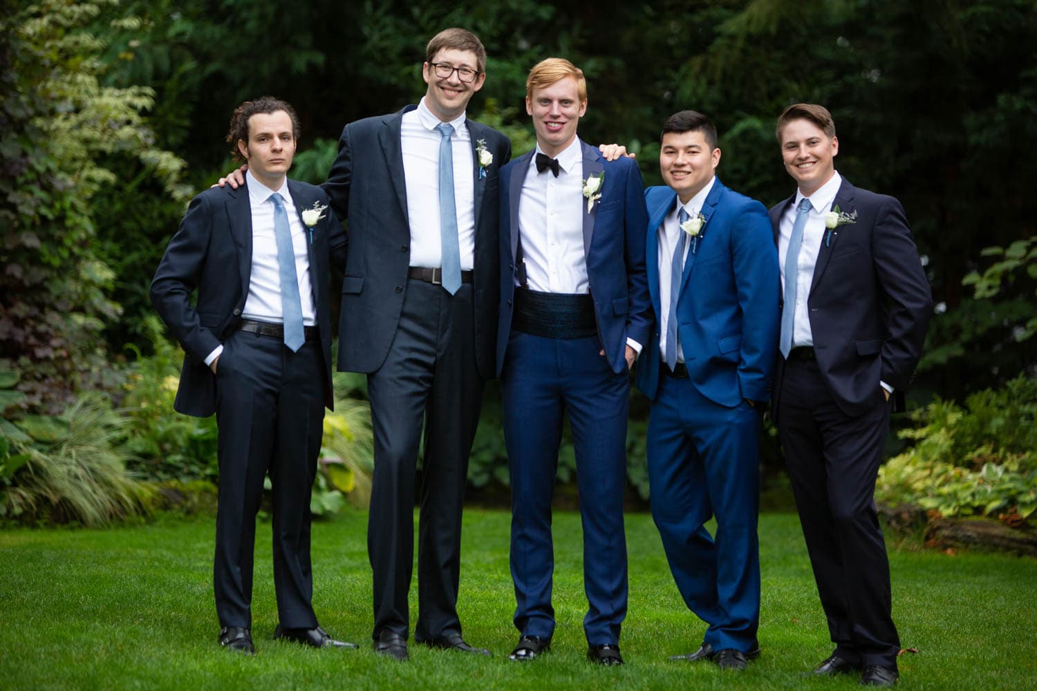 A groom in a bow tie and cummerbund in a navy suit poses with his groomsmen, in navy suits with white shirts and light blue ties.