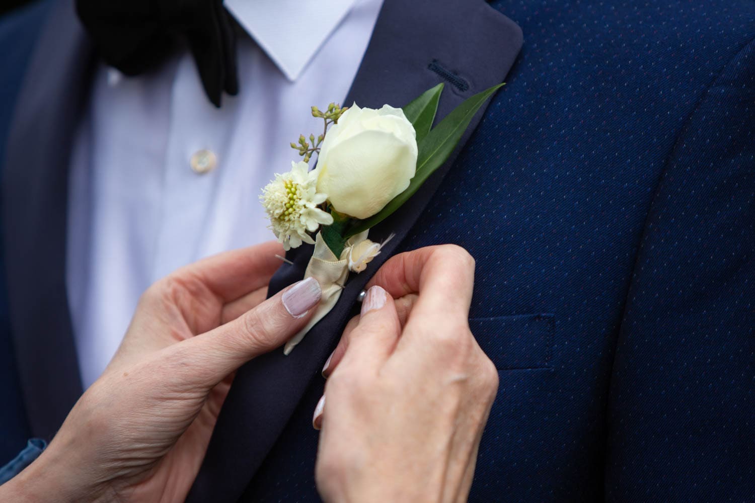A close up photo of the mother of the groom pinning the boutonnière to her son's lapel. It has a white rose, a white mini dahlia, two tree leaves, and ivory ribbon.