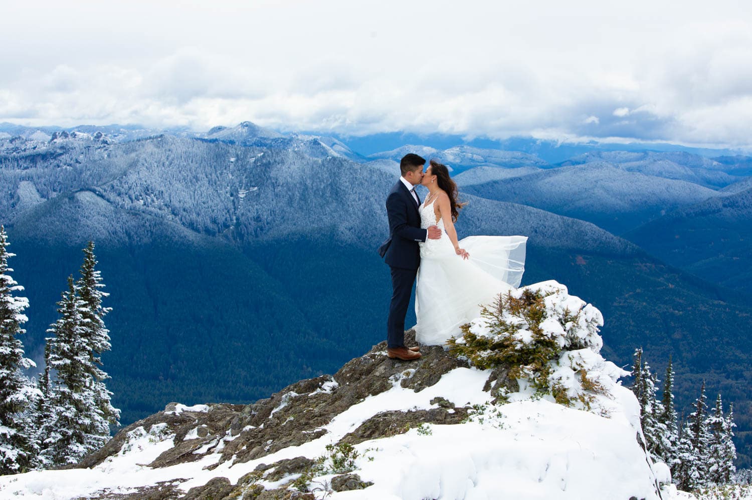 With the wind dramatically blowing the bride's hair and dress back behind her, the couple kiss on a rock promontory high in the mountains near Mt. Rainier with rolling snow-covered hills behind them and puffy clouds filling the sky.