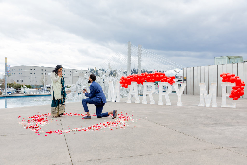 A young Black man with a beard and a short afro in a blue suit proposes on one knee to his girlfriend in front of large letters that spell "MARRY ME." They are standing inside pink and red rose petals in the shape of a giant heart. Behind them is the outdoor glass art Fluent Steps by Martin Blank at the Museum of Glass, the Tacoma Dome, and the East 21st Street Bridge in Tacoma, WA.