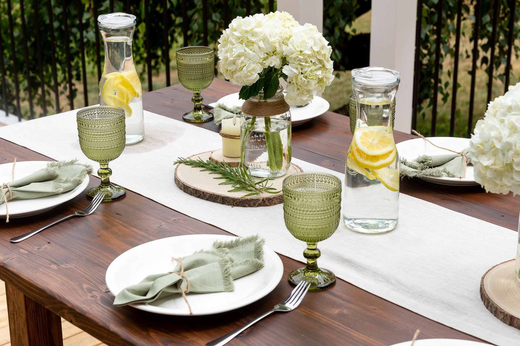 Vase with white hydrangeas, lemon water pitchers, and green glasses on a rustic outdoor dining table.