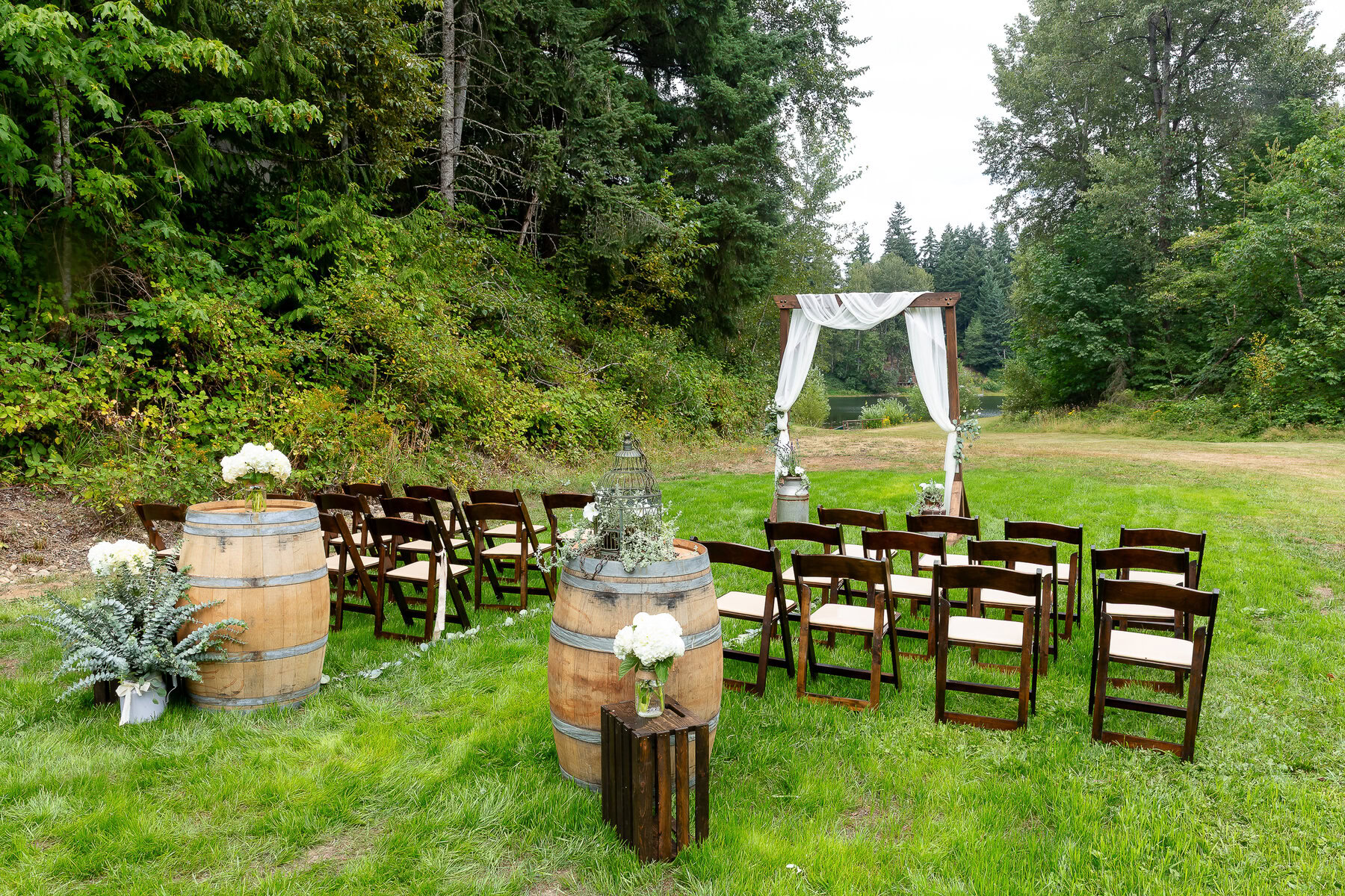 Rustic outdoor wedding ceremony setup with wooden chairs, floral decor, and draped arbor in lush garden setting