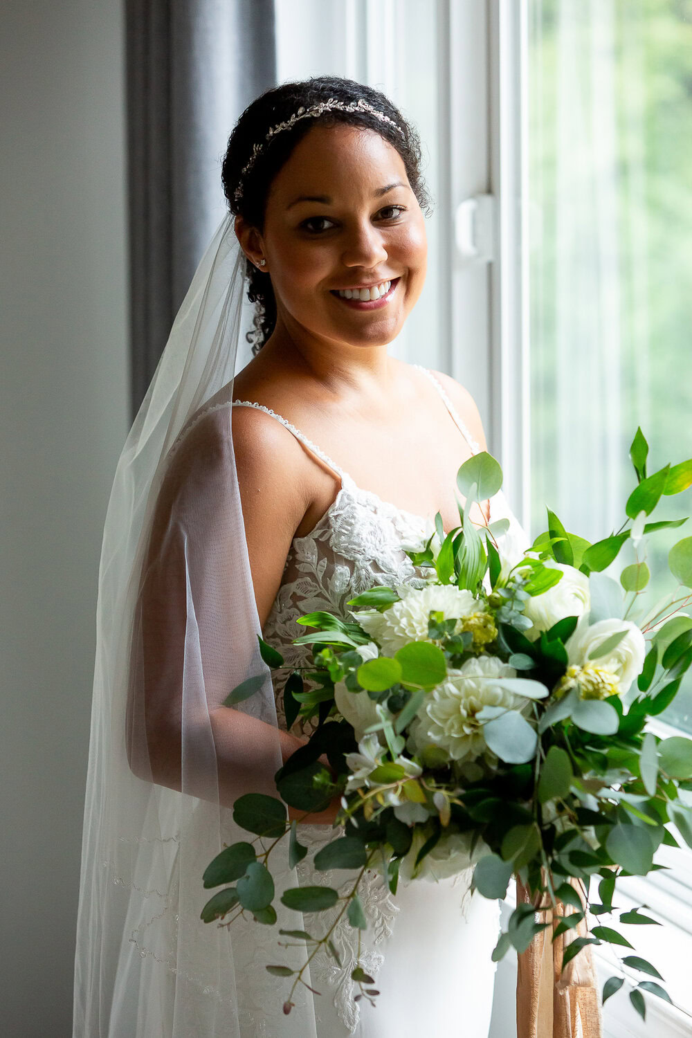 Elegant bride holding a lush white and green bouquet, soft natural light from window, wedding photography by Nallayer Studios.