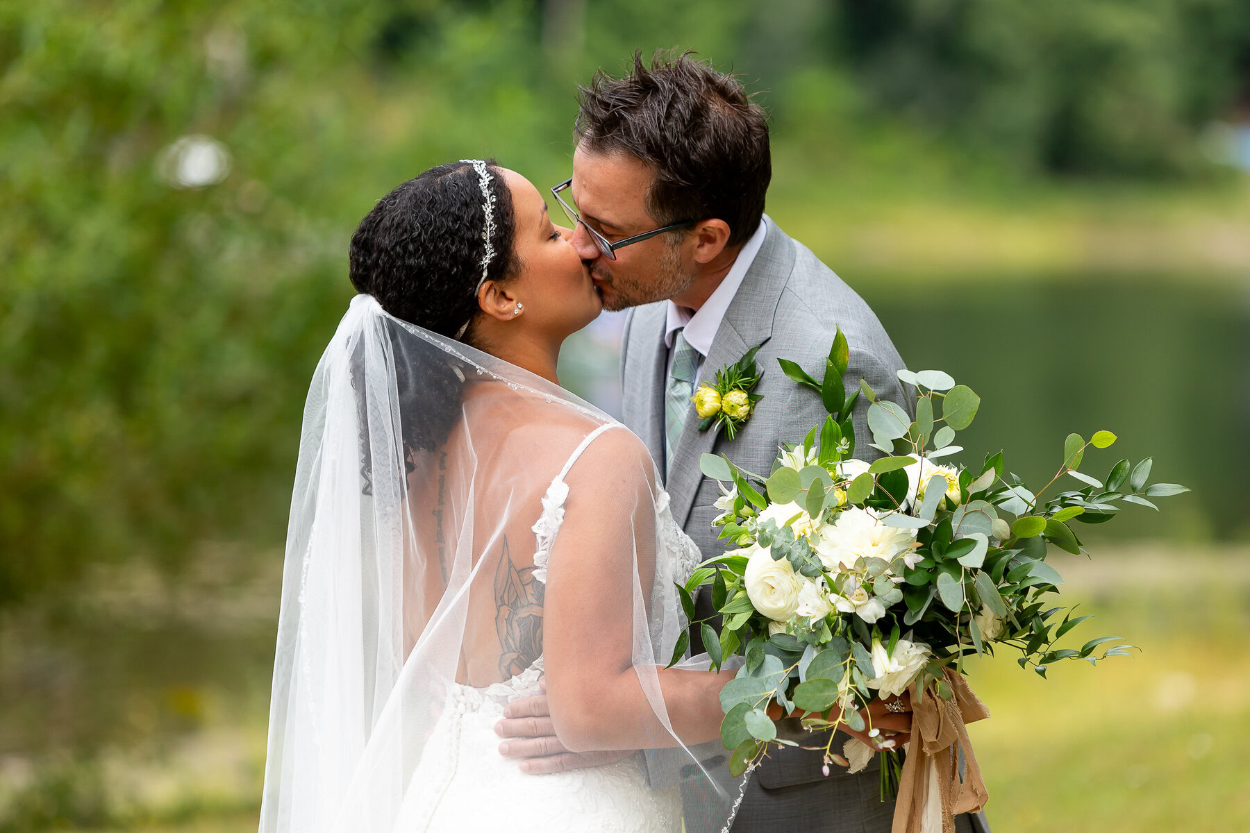 Beautiful wedding couple sharing a romantic kiss outdoors, holding a lush flower bouquet, natural greenery background.