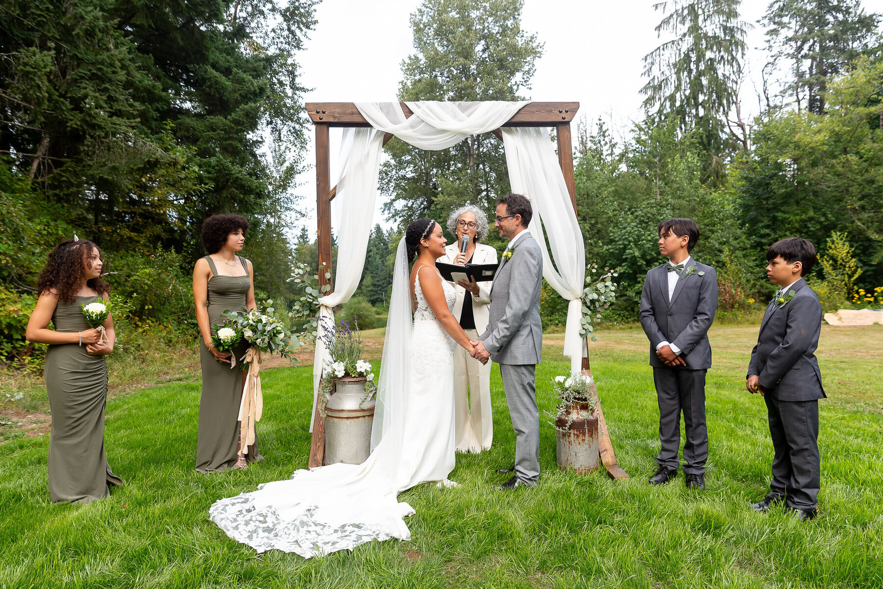 Elegant outdoor wedding ceremony with bridal party under a stylish wooden arch in lush greenery.