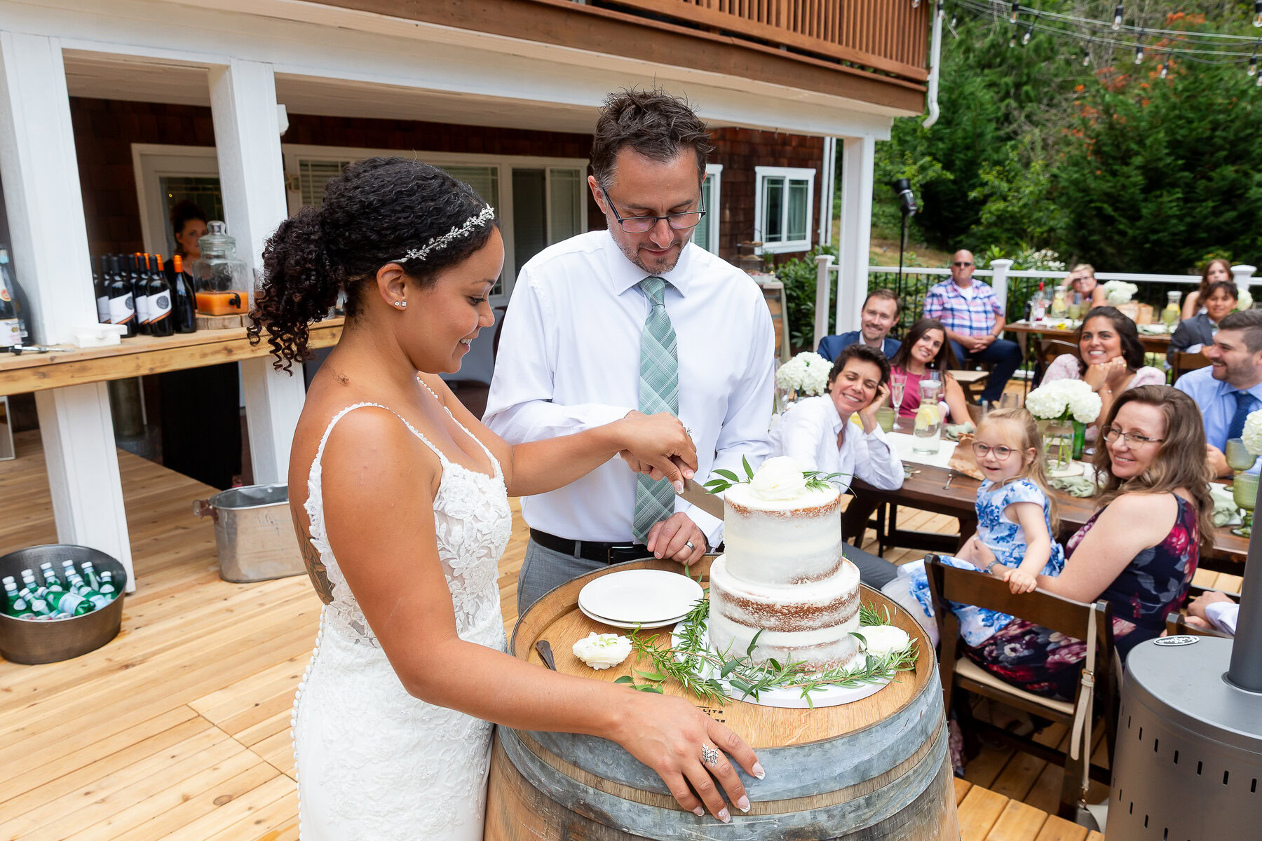 Elegant bride and groom cutting wedding cake at outdoor reception with guests smiling in the background