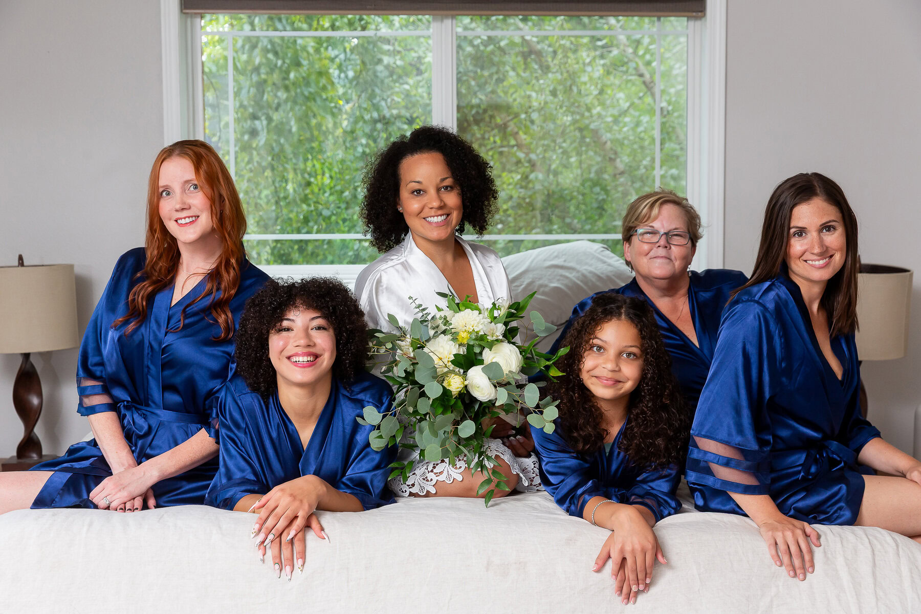 Elegant women’s wedding party in matching blue satin robes, celebrating with bouquet, indoor in front of large window.